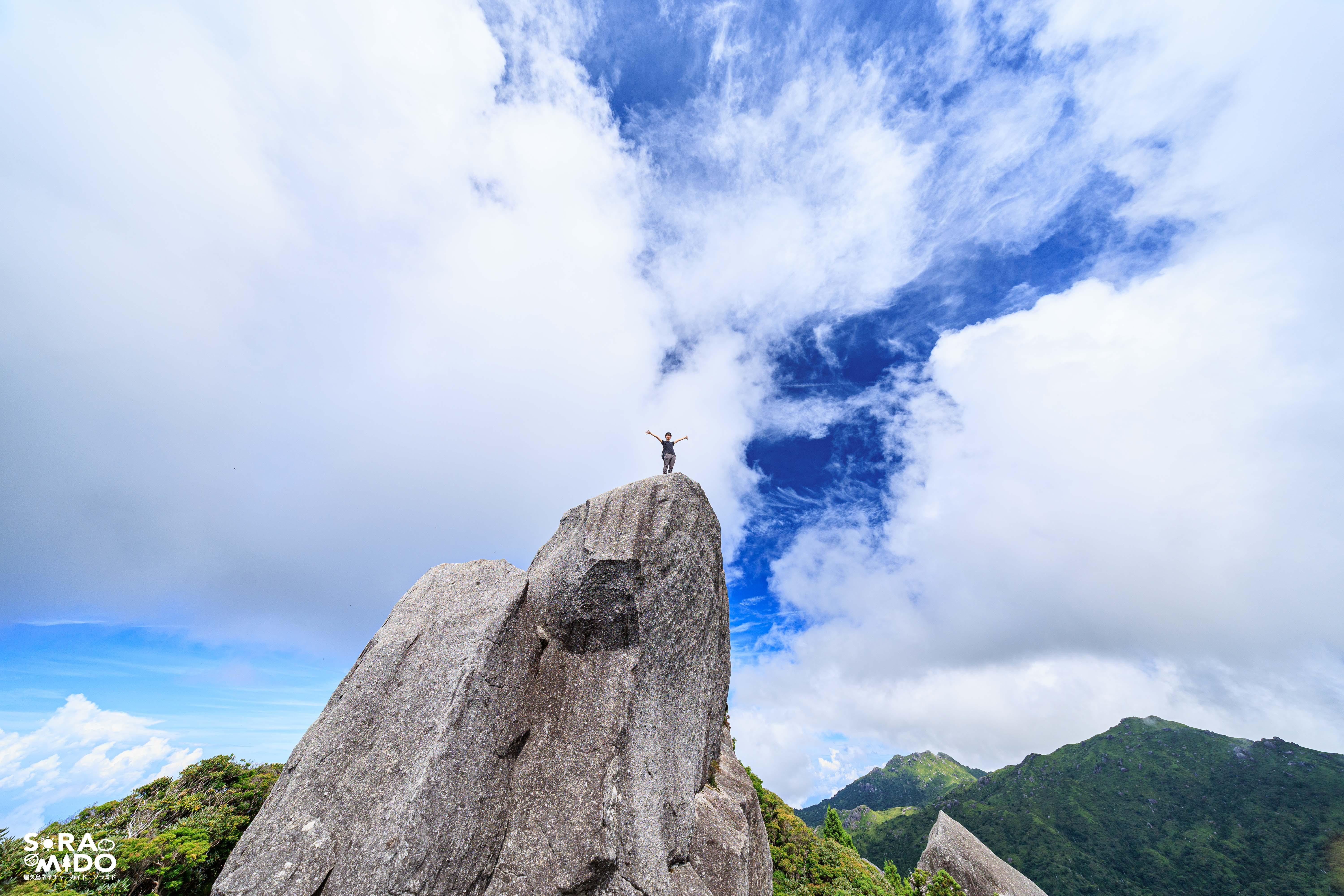 夏の青空の下、黒味岳の頂上で両手を広げて立つハイカー。