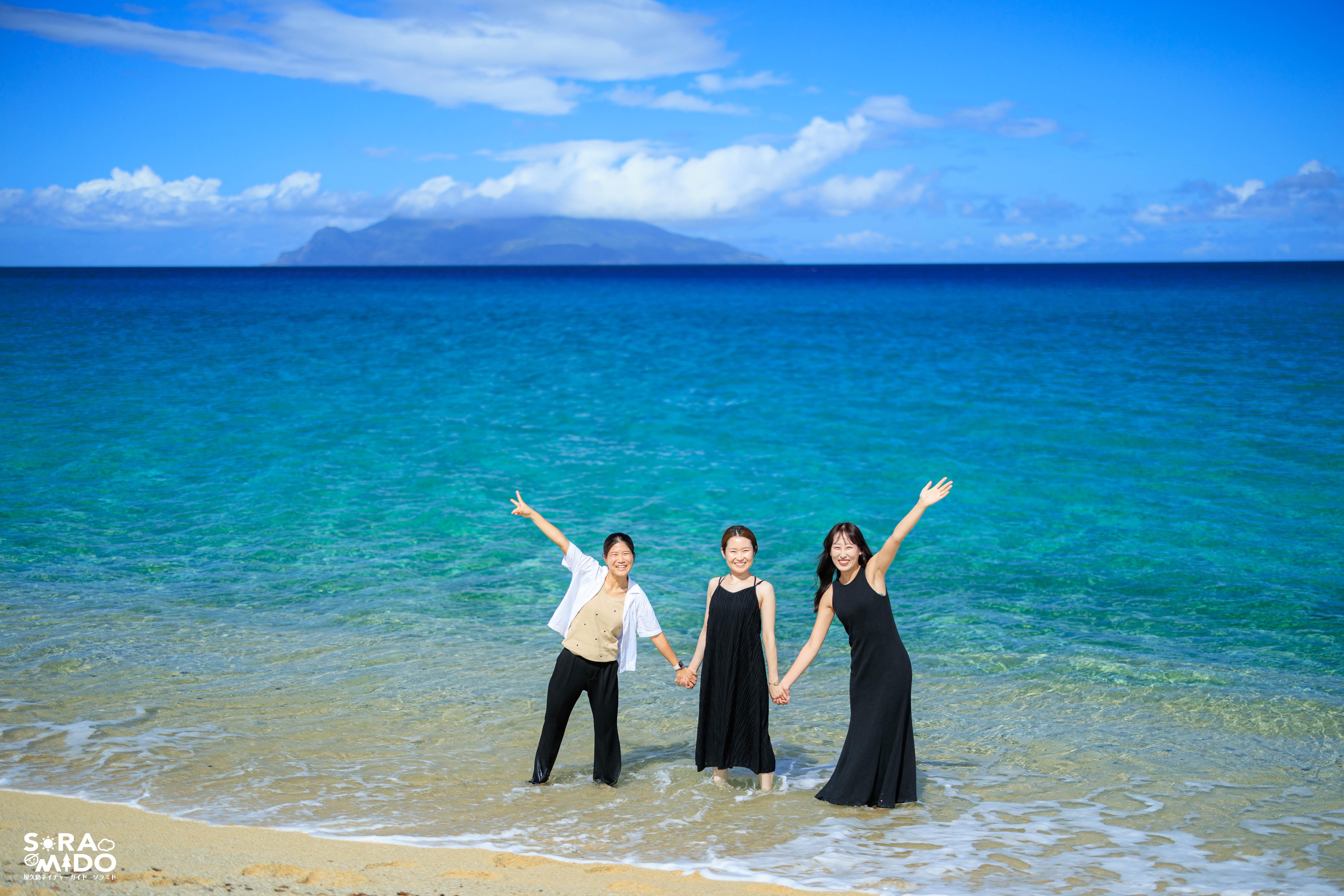 透き通るスカイブルーの海を背に、手をつないで笑顔ではしゃぐ３人の女性旅仲間