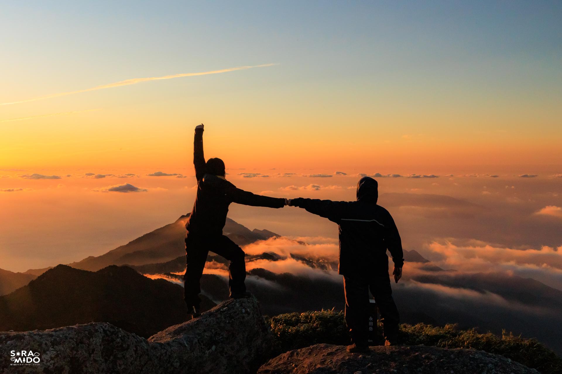 雲海に浮かぶ夕日の中、永田岳山頂で手を取り合い達成感を分かち合う登山者たち！