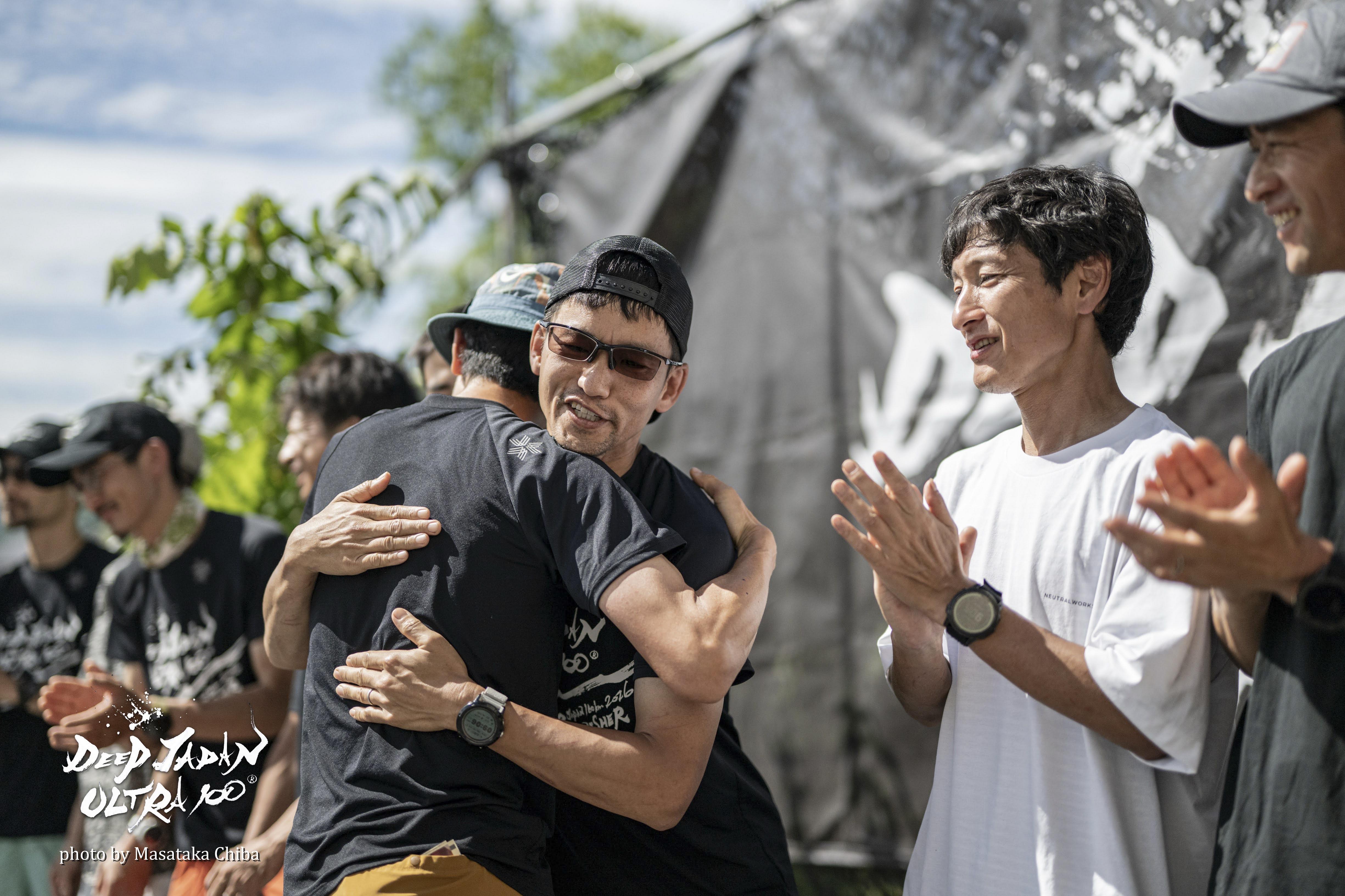 DEEP JAPAN ULTRA 100の表彰式で、5位入賞した仲間の大木さんと抱き合って健闘を称え合う様子。周囲の選手たちから拍手が送られている。